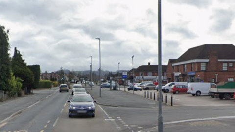 A street with cars driving along and parked beside a row of shops and buildings, with a car park under a cloudy sky.