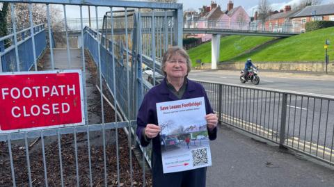 Campaigner Debbie Hills next to the closed footpath with Leazes Footbridge in the distance. Ms Hills wears a purple coat and glasses and holds a Save Leazes Footbridge poster. A motorcyclist is travelling on the road.