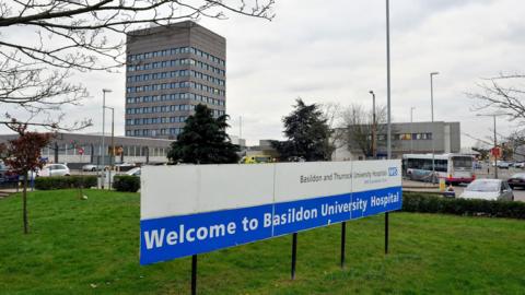 A white and blue sign at the entrance to Basildon Hospital, welcoming people to the site. In the background is a tall hospital building which looks similar to a block of flats, as well as several other grey buildings.