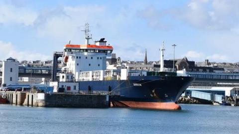 The MV Arrow moored in Douglas Harbour alongside the Victoria Pier. It is navy blue, white and orange. The sea is blue and there is blue sky above that is partially cloudy.