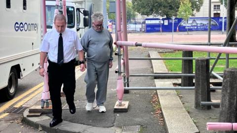 Anthonio Villafane is wearing a grey tracksuit with white trainers. He has grey/white curly hair and black glasses. He looks down as he is led by a Geoamey court officer who is wearing a white shirt, black trousers, black boots and a black tie. Anthony Villafane is handcuffed to the court officer by the wrist.