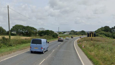 Cars travelling along a road. Grass verges are on either side of the road. The cars are driving in single file.