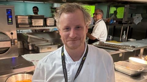 Jon Batchelor is wearing chef's whites and is standing inside a kitchen. He is looking directly at the camera and smiling.