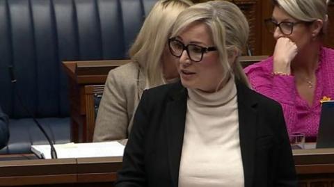 Michelle O'Neill has blonde hair, tied in a pony tail and glasses. She is wearing a white polo neck jumper and a dark jacket