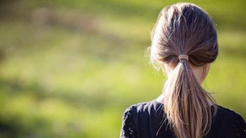 A girl with a ponytail looking away from the camera, outside in a green space.