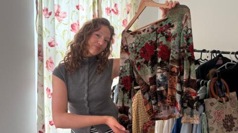 A young woman with curly red hair holding up a shirt and smiling. She is wearing a grey top and in the background is a rack of various pieces of clothing