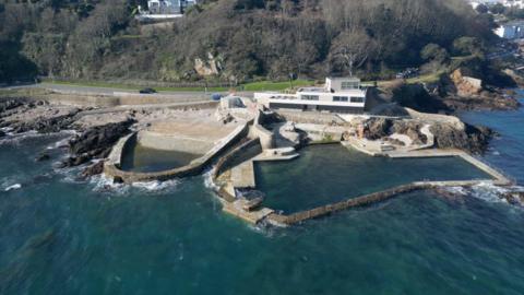 Aerial view of La Vallette bathing pools in Guernsey, showing stone sea walls, a slipway and a rectangular tidal pool beside a coastal road and buildings above rocky shoreline.
