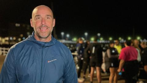 Peter is in a blue Montane jacket smiling at the camera in the foreground, with a group of people gathered in the brightly lit background at night near a waterfront promenade