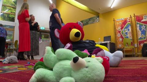 A pile of brightly coloured teddy bears laid on the floor of a children's centre. There are three people in the background talking. 