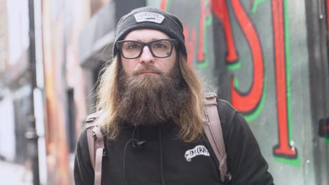 Tom is looking towards the camera. He is wearing a black hoodie, black beanie and black glasses. He is also wearing a light pinkish backpack. 