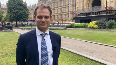 Hamish Falconer standing outside the Houses of Parliament dressed in a blue suit 