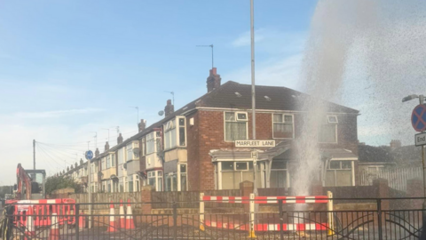 Water can be seen gushing out of a water main on Marfleet Lane, the leak is behind orange barriers and a sign on a lamppost reads Marfleet Lane. There are houses in the background.