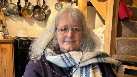 Penny Bayer, wearing a blue zip up jumper and blue and white scarf. She has shoulder length grey hair and is wearing glassed. She is sat down facing directly at the camera. Behind her to the left is a cabinet and a cooker, on the wall there are pots and pans on hooks. To the right behind her are stairs.