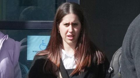 A woman with dark hair, wearing a black suit and white shirt. She is standing outside a court. 