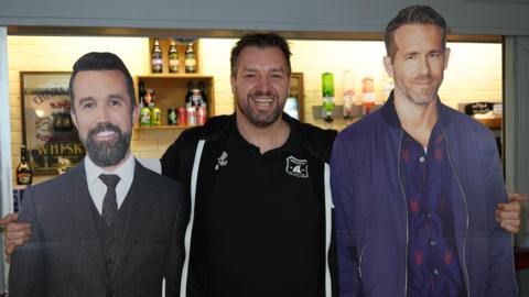 Wroxham FC chairman James Blower wearing a black hooded top and standing with his arms around two cardboard cut outs of actors Ryan Reynolds and Rob McElhenney