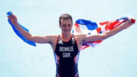 Alistair Brownlee holds two flags: the Union Jack and Yorkshire's white rose flag after winning gold in the Rio OIympics. He is a man with short dark hair wearing a black and red vest. 