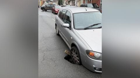 A silver car has its front driver's side wheel stuck in a sinkhole, which has appeared on a residential road. There is queueing traffic behind the car, and an SUV-type car can be seen turning around.