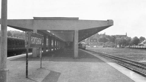 Old archive picture in black and white of old railway station. There's a big sign saying Portishead indicating the stations names. There's a platform with several trains on the track with trees and a church in the distance.