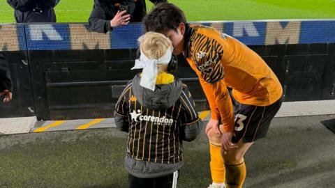 A footballer wearing a distinctive orange Hull City tiger striped kit speaks closely to a young child with bright blond hair. The little boy wears a white bandana and ear defenders.