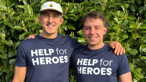 Two young men standing in front of some foliage, with their arms around each other. The one on the left is taller and wearing a white baseball cap, while the other has brown hair. Both are wearing dark-blue t-shirts with Help for Heroes written on them in white letters.

