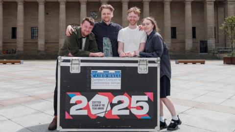 Three young men and a woman stand outside a large hall in a city square. They are leaning on a large packing case with the words Scottish Album of the Year 2025.