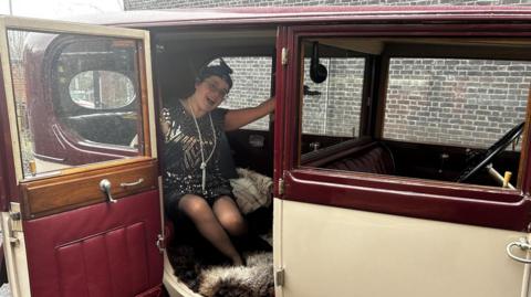 A woman in a vintage car wearing a costume which looks to be 1920s-themed.