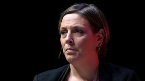 Jess Phillips has short brown hair and is sitting at an angle against a black background. Her head and shoulders are visible, and she is wearing a black suit jacket