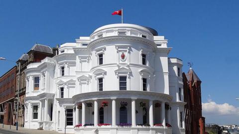 The exterior of the Tynwald legislative buildings, a grand round white structure with colums at the bottom, a Manx glag flies at the top, on a clear blue day.