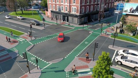 A computer generated image showing a junction in a city centre. There are four roads at the traffic light junction, with a bus lane between two buildings at the top and the other three roads holding regular traffic. Green cycle lanes border the central part of the junction, which a red car is making its way across, and pedestrians can be seen waiting at the lights to cross.