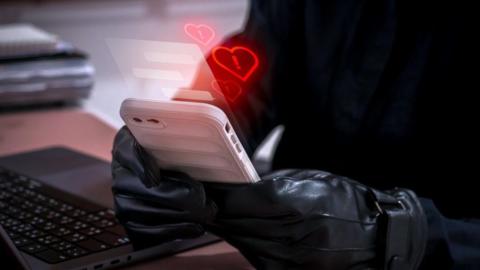 Romance scam concept showing a cybercriminal wearing black gloves using a white smartphone to deceive victims with fake love messages with red hearts with exclamation marks coming from the phone. A laptop is next to them on a desk.