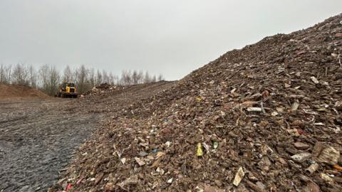 A close up image of a large pile of waste which extends out into the distance of the photograph. The waste is made up of wood, plastic and other bits of household waste. There is a yellow JCB truck in the background. 