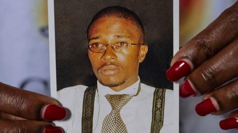 A close-up of a woman's hands, with read nail varnish, holding up a small photo of Floribert Bwana Chui Bin Kositi. In it he is staring ahead and looking serious. He is wearing oval wire-rimmed glassed, a silver and black dog tooth design tie, black braces and a white shirt.