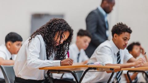 An exam hall of students, one female student is the focus of the picture, she is concentrating on her paper and writing with a red pen. Behind her is a male pupil reading the paper. Other students in the background, as well as a teacher, are out of focus but you can tell from their body language they're struggling.