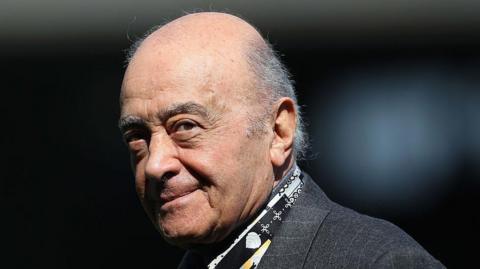 A close up on Mohamed Al Fayed, who is wearing a grey suit and has a bald head. The background is dark and blurred and his head it tilted so he is looking at the camera.