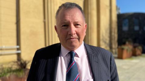 A man with short grey hair looking at the camera. He is wearing a light pink shirt with a navy tie that has light blue and red stripes. He has a pinstripe blazer on.