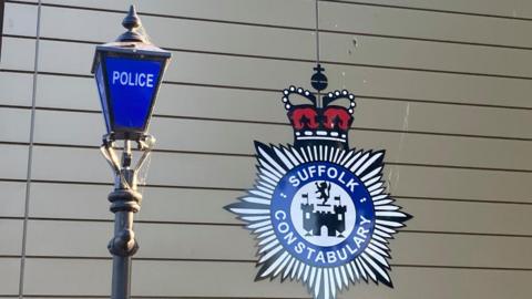 A Suffolk Constabulary badge on a wall with a blue police lamp-post in front of it. 