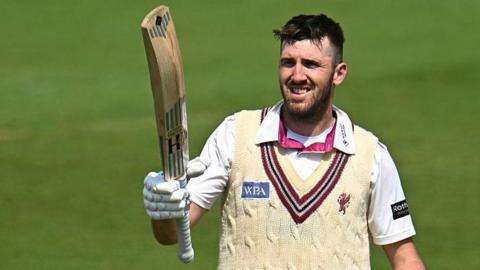 Craig Overton holds a bat towards the sky in his right arm, as he stands on the pitch during a match for Somerset