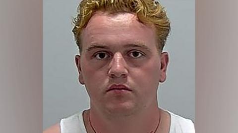 A police custody mugshot of Johnny Ward. He is wearing a white vest and a necklace.