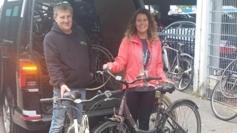 A man and a woman holding rusty bikes. They are standing in front of a large black vehicle. There are other bicycles in the background