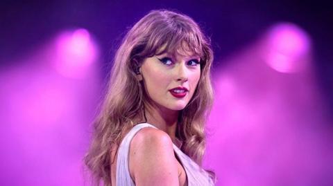 A close-up photo of Taylor Swift on stage. She has blonde wavy hair and wears a light purple dress.