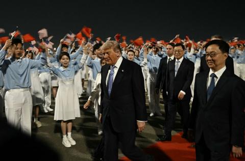 A man wearing a dark suit walks past a row of people wearing blue and white and waving flags on a red carpet