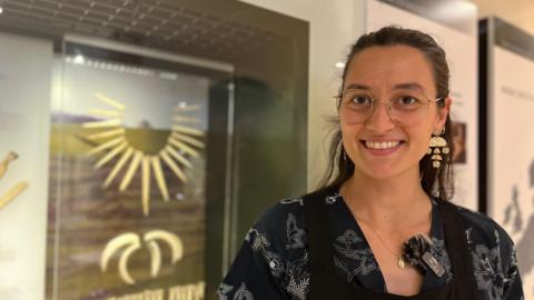 Amber smiles at the camera. She stands in front of a blurred cabinet with items in.
She wears glasses and black floral top.