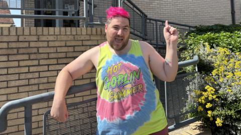 Aaron Weight stands in front of the BBC Radio Suffolk building, he is dressed in a blue, pink and yellow tank top. His hair is bright pink and he is smiling while pointing up to the sky.