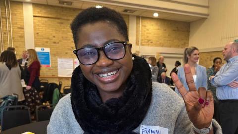A woman with glasses smiles, and gives the peace sign with the backdrop of the hackathon event. 