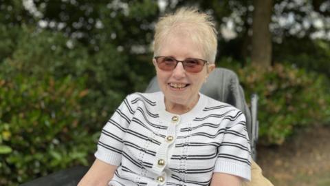 A woman with short blonde hair is smiling and sat in a chair outside with shrubbery and trees behind her. She is wearing a white and black stripey top and tinted glasses.