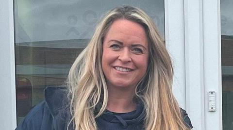 Close up shot of Nicky Morson smiling. She has long blonde hair and is standing in front of a window. She is wearing a black jacket. 
