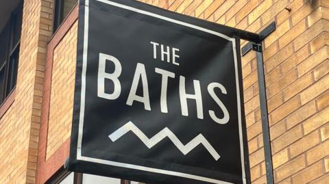 The Ipswich Baths sign above the building. It shows a black sign with 'The Baths' written in write writing. The sign is overhanging from the side of the building. 