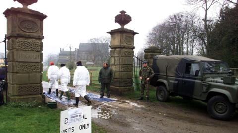 A soldier guards the entrance to a farm as three people in white hazmat suits walk onto the premesis.