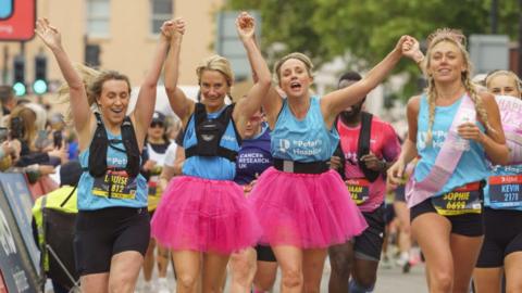 Three young women smile and link hands as they run in the Bristol Half Marathon. Two of them are wearing pink skirts below their running gear