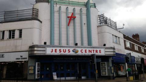 A large white and green building with "Jesus Centre" written across it with a large red crucifix on the outside.